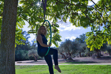 Young blonde woman in a blue suit, sitting on an aero hoop hanging from a tree in the middle of a park. Concept aerial hoop, pole dance, fitness, curvy girl.