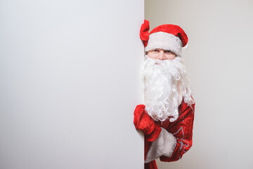 Santa Claus in a traditional red costume with beard peeks out from behind a white background.