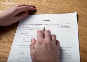 Obraz premium Paper contract on wood desk and female hand with pen studying agreement.