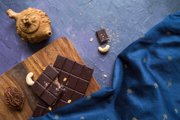 Top view of vegan chocolate bar on wooden cutting board with cashews and chinese style tea pot on blue painted background draped with table cloth