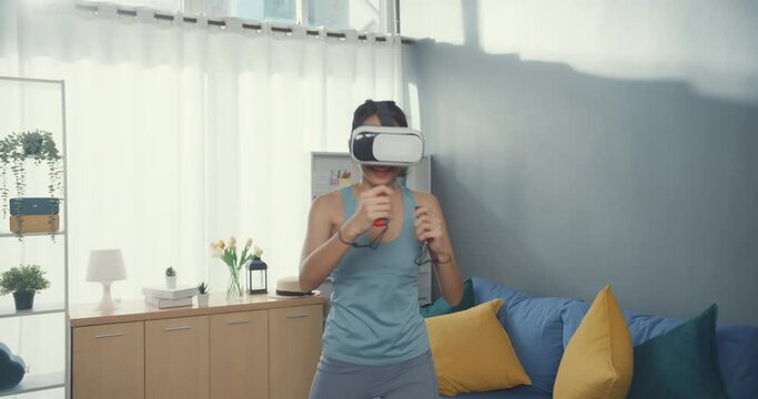 Young attractive Asian woman with sportswear wearing virtual reality headset play switch game boxing exercise online program in living room at house. Home quarantine workout and fitness concept.