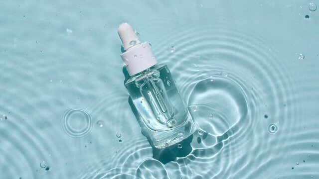 Glass cosmetic bottle with pipette lies on the surface of the water. Top view of water splash blue colored. Pure blue water with reflections sunlight. Sample of packaging for design, advertising.4k.