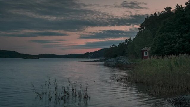 Time lapse of Swedish red hut at lake in Sweden during sunset, moving clouds in the sky, reed in the front
