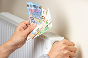 Woman hand holding euro banknotes and adjusting temperature of central heating radiator at home....