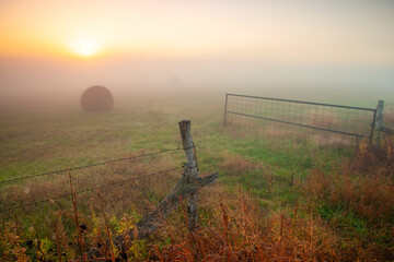 Open gate entrance into grassy field with hay bale at sunrise with morning ground fog © Lenspiration