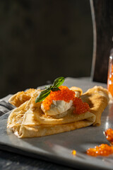 Crepes (big thin pancakes) with red caviar, and cream cheese or sour cream with basil leaf on dark background. Beautiful light, selective focus, copy space. Tasty breakfast, dark mood photo concept.