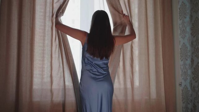 Young woman approaches window, pushes aside curtains and white tulle with hands, opens window and enjoys fresh breeze. She has dark hair and wearing blue shirt. Morning, rest, the beginning of new day