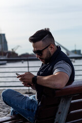 Obraz premium young man with sunglasses and beard siting by a lake in winter using his cellphone