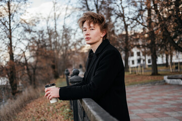 Thinking young man in black coat standing with cup of coffee in hands on a city street leaning on fence and looking away. Handsome man waiting for someone. Breathing fresh air in autumn park.