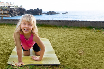 Cute happy little girl in sportswear smiling at camera while while exercising on seaside