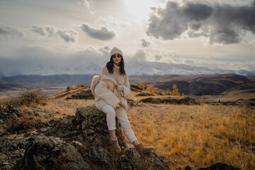 Fototapeta premium Beautiful stylish girl tourist posing against the backdrop of white snow peaks. Black glasses, white hat, white jeans, white down jacket. Sunny. Altai Severo Chuisky Ridge. High quality photo