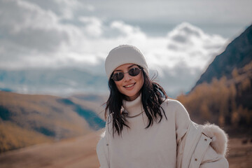 Beautiful stylish girl tourist posing against the backdrop of white snow peaks. Black glasses, white hat, white jeans, white down jacket. Sunny. Altai Severo Chuisky Ridge. High quality photo