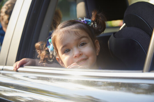 Little toddler girl looking out the car window with pig tails, big smile and sunshine glaring off vehicle