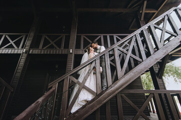 Beautiful, stylish newlyweds hug and kiss while standing on a wooden staircase in a big house. Wedding portrait, photography.