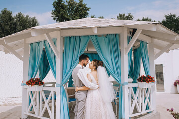 A young, bearded groom in a gray suit and a beautiful bride in a white dress are hugging against the background of a wooden gazebo with curtains in Turkey against the background of nature, the sky.