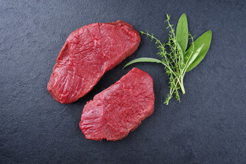 Raw dry aged bison beef rump steak slices with herbs offered as top view on black background with copy space