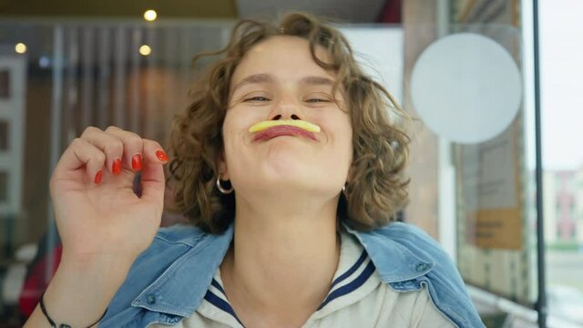 Funny and cute happy young woman giggle and smile, laugh into the camera with real and authentic emotions, play with her food, hold french potato fry under nose like moustache. Candid teenager