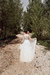 view from back. bride in wedding dress walks along mountain path between trees.