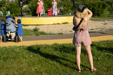 A girl in Shorts and a top stands in a city park next to the stage, on her head a gray bandana, a view from the back. Solo day, city holiday.