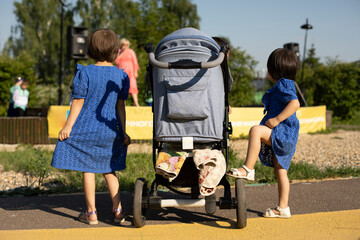 Two children next to the stroller on a walk on a sunny day. Girls in blue dresses, a view from the back. - obrazy, fototapety, plakaty