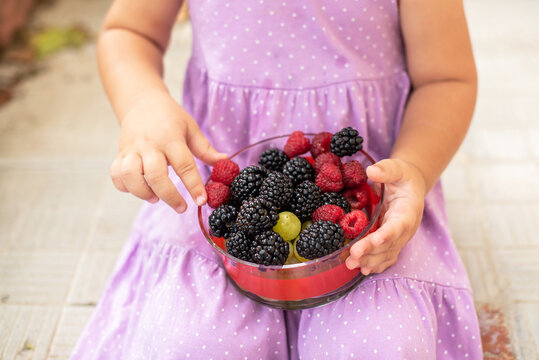 Little cute girl happy to eat fruits - raspberries and blackberries. Caucasian Toddler holding bawl with fruits outdoors. Yummy and healthy food for kids. Healthy nutrition,  eco food