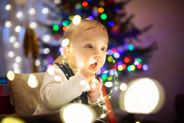 Happy baby boy playing with Chistmas lights in a cozy living room on Christmas eve. Celebrating Xmas at home. Winter evening with family and kids.