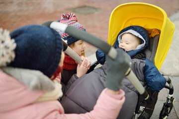 Two older sisters admiring their little brother sitting in a stroller outdoors. Little child in pram. Infant kid in pushchair. Winter walks with kids.