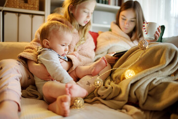 Two young sisters and their baby brother snuggling up on the sofa in a cozy living room at Christmas. Cute children using a tablet at home during winter break.