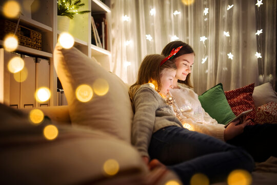 Two cute young sisters using a tablet pc at home in warm and cozy living room at Christmas. Family having online video call on Xmas eve.
