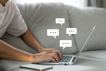 A young asian man wearing a white t-shirt sitting on sofa in living room using a laptop hand touching keyboard with a chat icon show that he is interacting with close friends or talking about work.