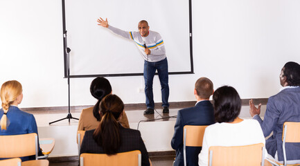 Excited Hispanic preacher giving motivational speech to business people from stage in conference hall