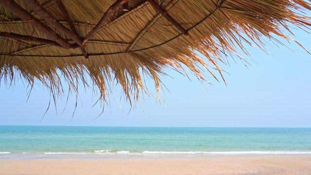 Thatched Roof Made Of Palm Leaves Provides Shade On Wide Sandy Beach With Slow Motion Little Waves Rolling Onto The Shore. Protection From The Sun. Luxury Island Resort Vacation Tropical Destination.