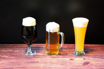 Three glasses of different shapes with different varieties of light, wheat and dark beers stand in a row on a wooden table.