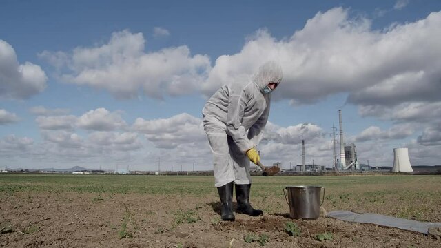 Laboratory assistant in protective workwear digging ground with shovel and bucket for soil sampling