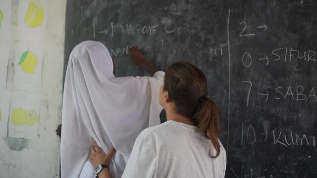 White caucasian teacher helping to black african girl whit white headscarf to write on the blackboard. Beautiful volunteer cinematic shot. Africa 4K.