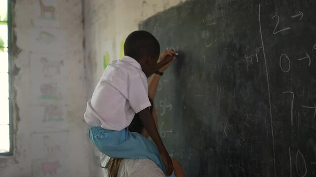 White caucasian teacher with black African child lifted on her shoulders helping him to write on the blackboard holding hands. Beautiful volunteer cinematic shot. Africa 4K.