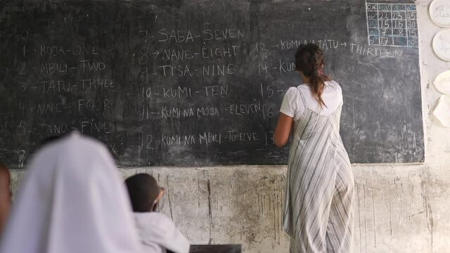 White Caucasian Teacher Writing On The Blackboard Numbers Teaching Black African Children. NGO Cooperation Volunteering Helping For The Development Of Education In Africa. Cinematic Shot 4K.