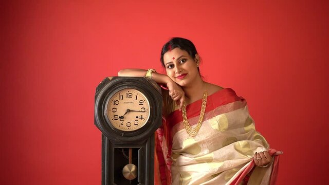 Kolkata : Traditionally dressed Indian female in red and white saree with a traditional vintage clock waiting in a red background studio.