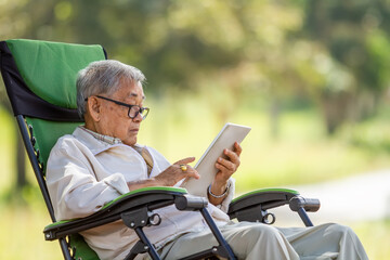 Asian senior man using digital tablet on folding chairat natural park. The use of technology by the elderly.