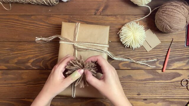 Person's hands ties gift box in craft paper with twine. Yarn and pom-poms on wooden table. Christmas decorations. Top view of festive composition.