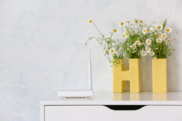 Modern wi-fi router and chamomile flowers on table near light wall