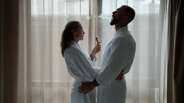 lovers are smoking e-cigarette in hotel room, man and woman are wearing white robes