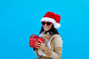 Beautiful young woman in Santa Claus hat in beige coat beret and sunglasses hold red gift over blue...