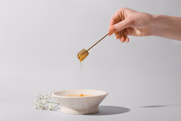 Woman pouring honey from dipper into bowl on grey background