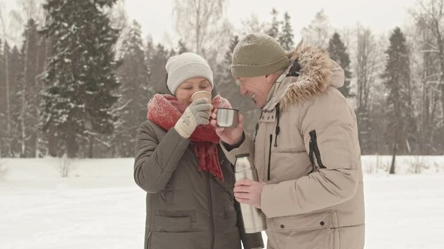 Medium of senior Asian woman and her Caucasian husband standing in park on snowy winter day, talking, smiling and drinking hot tea