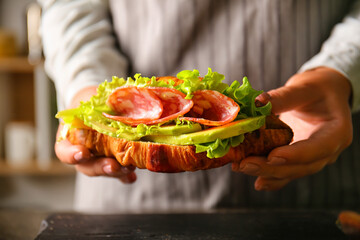 Woman holding tasty croissant sandwich with meat at kitchen table