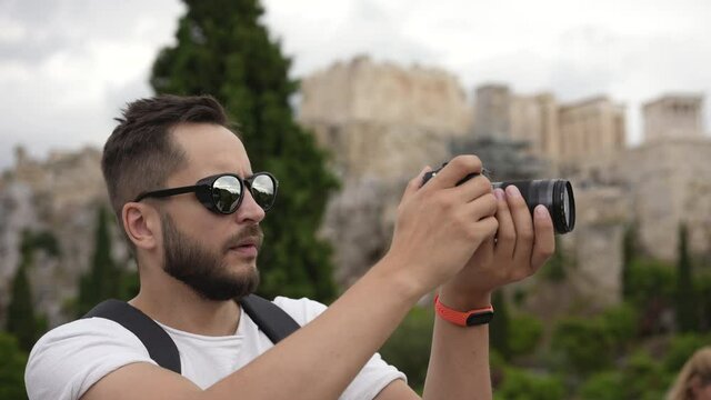 Young adult tourist stands on hill with camera, spends time on taking pictures of historical Greek landmark. Traveler on sight of Acropolis in Athens. Tourism, traveling to ruins of old civilization.
