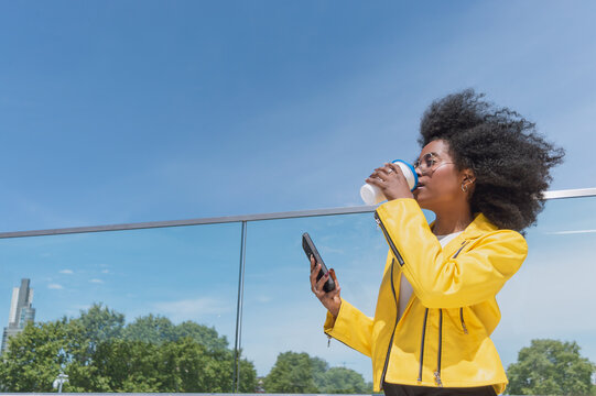 young black hispanic latina woman with afro outside looking to the right, drinking coffee and holding her phone in her hand