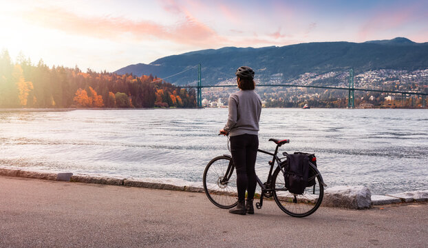 White Caucasian Adult Woman riding a bicycle on Seawall in Stanley Park. Sunset Sky Art Render. Downtown Vancouver, British Columbia, Canada.