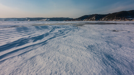 Frozen Lake Baikal. The snow on the surface forms wavy patterns similar to dunes. A mountain range against a blue sky.